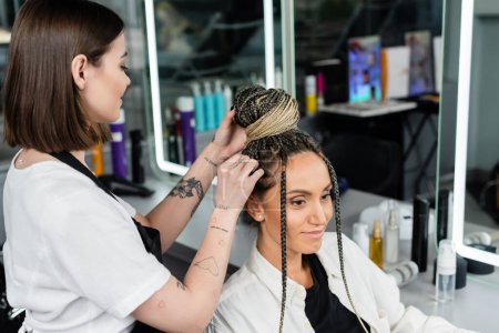 Photo for Salon experience, tattooed hairdresser doing hair bun to female client with braids, happy women, client satisfaction, customer in salon, beauty service, feminine, hair make over - Royalty Free Image