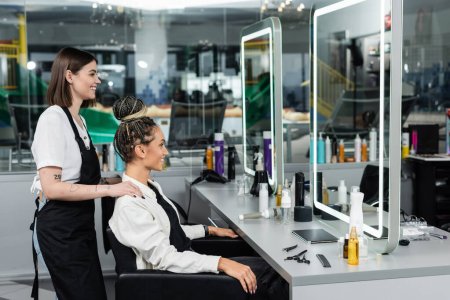 Photo for Client satisfaction, salon experience, happy hair stylist and female client with braids looking at mirror, cheerful women, hair bun, customer in salon, beauty service, tattooed, side view - Royalty Free Image