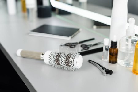 Photo for Salon accessories, focus on round brush and professional hairdressing hair clip, hair cutting tools, hairdressing scissors, bottles, hair oil near mirror in beauty salon, beauty industry - Royalty Free Image