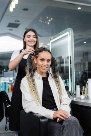 Photo for Hairdresser and client, beauty salon, cheerful woman with braids and two ponytails smiling near hair stylist in salon, customer satisfaction, beauty worker, professional, hair fashion - Royalty Free Image