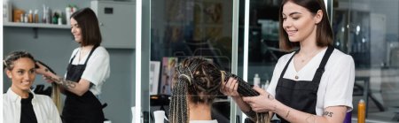 beauty worker and client, beauty salon, tattooed hair stylist doing hair of cheerful woman with braids, two ponytails, customer satisfaction, beauty worker, hair fashion, mirror reflection, banner