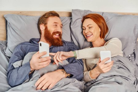 Photo for Screen time, tattooed couple using smartphones, networking, relaxing on weekends without kids, husband and wife, redhead woman pointing at mobile phone of bearded man, cozy bedroom - Royalty Free Image