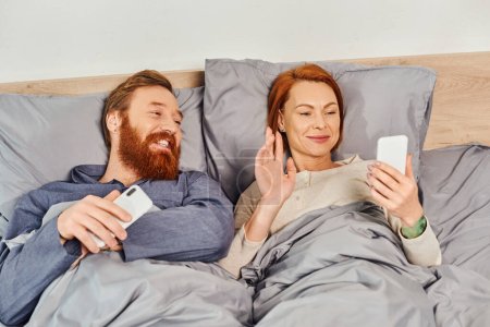 screen time, tattooed couple using smartphones, relaxing on weekends without kids, husband and wife, redhead woman having video call near bearded man, cozy bedroom, waving hand