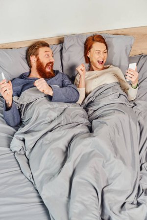 Photo for Tattooed couple using smartphones, networking, relaxing on weekends without kids, excited husband and wife, bearded man and redhead woman with mobile phones, cozy bedroom, screen time - Royalty Free Image