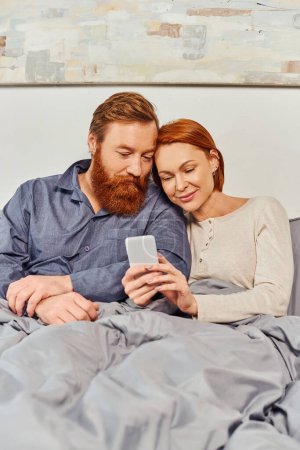 digital couple, screen time, networking, relaxing on weekends without kids, pleased husband and wife, bearded man and redhead woman using smartphone, tattooed, day off 