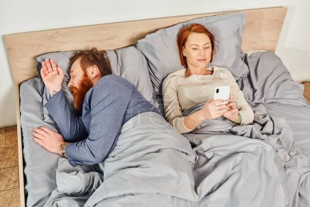 Photo for Quiet house, parents alone at home, redhead husband and wife in cozy bedroom, bearded man sleeping near carefree woman using smartphone, networking, screen time, day off, tattooed - Royalty Free Image