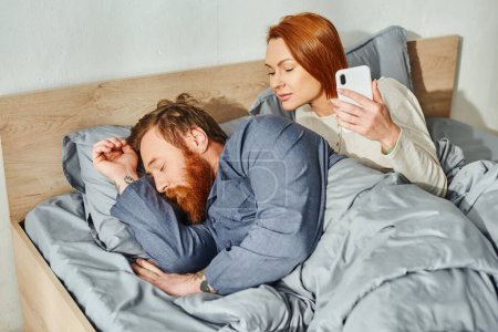 quiet house, parents alone at home, redhead wife looking at husband, bearded man sleeping near woman using smartphone, networking, day off, wake up, tattooed, couple without kids 