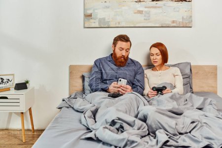 quiet house, couple without kids, redhead husband and wife, bearded man using smartphone near woman holding joystick, networking, playing video game, day off, tattooed, parents alone at home 