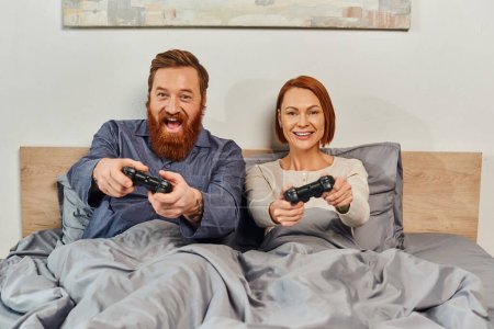 weekends without kids, redhead husband and wife playing video game, bearded man and happy woman holding joysticks, excited, gaming fun, married couple, modern lifestyle 