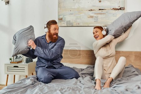 Photo for Day off without kids, having fun, shared music, redhead husband and wife, happy couple in wireless headphones pillow fight, bearded man and carefree woman, tattooed music lovers, weekends - Royalty Free Image
