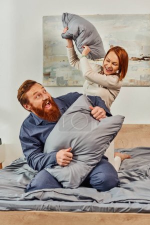 parents alone, having fun, pillow fight, redhead husband and wife, happy couple in sleepwear enjoying time together, bearded man and carefree woman, tattooed people, day off without kids