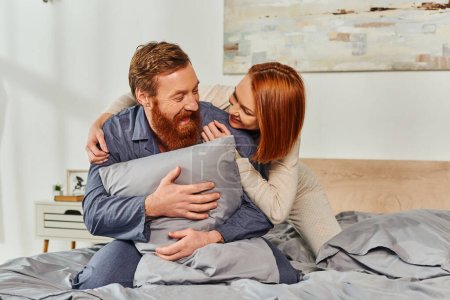 Photo for Day off without kids, redhead woman hugging bearded husband with pillow, happy couple in sleepwear enjoying time together, tattooed people, parents alone at home, quality time together - Royalty Free Image