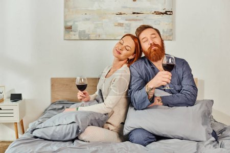 Photo for Day off without kids, married couple holding glasses of red wine, redhead husband and wife, enjoying time, day off, weekends together, pleased and tattooed, parents alone at home - Royalty Free Image