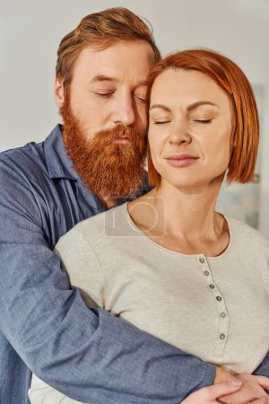 Photo for Married couple hugging in cozy bedroom, day off without kids, redhead husband and wife, enjoying time together, weekends together, tattooed, bonding, parents alone at home, closed eyes - Royalty Free Image