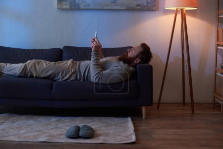 Photo for Digital age, mobile interaction, bearded man with red hair using smartphone, resting on sofa, painting on wall, slippers on carpet, night, light from lamp, leisure time, cozy living, side view - Royalty Free Image