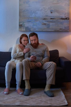 social media, night, redhead couple spending time without kids, day off, husband and wife, bearded man using smartphone near woman, comfortable living, cozy living room, carefree, tattooed 
