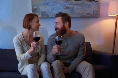 Photo for Quality time, couple without kids enjoying time at home, tattooed couple relaxing on weekends, holding glasses of red wine, husband and wife, bearded man and redhead woman, cozy living room, day off - Royalty Free Image
