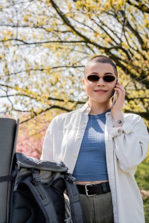 Lächelnde, kurzhaarige und tätowierte Reisende in Sonnenbrille und lässiger Kleidung, die mit dem Smartphone spricht, während sie in der Nähe des Rucksacks mit der Natur im Hintergrund steht, selbstbewusste Forscherin