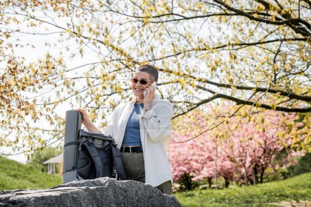 Lächelnde, kurzhaarige und tätowierte Touristin mit Sonnenbrille, die mit dem Smartphone spricht und in der Nähe eines Rucksacks auf einem Stein steht, mit malerischer Natur im Hintergrund, selbstbewusste Forscherin
