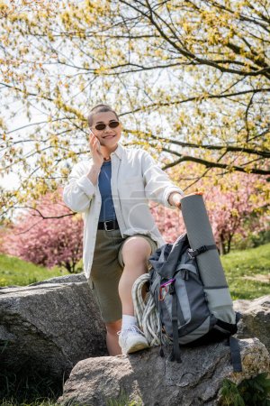 Jeune touriste aux cheveux courts positive en vêtements décontractés et lunettes de soleil parlant sur smartphone tout en se tenant près du sac à dos et des pierres avec la nature en arrière-plan, exploratrice confiante