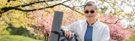 Positive young and short haired female backpacker in casual clothes and sunglasses standing near backpack with fitness mat with nature at background, confident female explorer, banner 