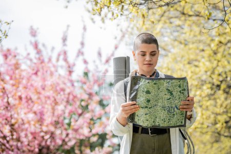 Young short haired female tourist in casual clothes with backpack looking at map while standing with blurred landscape at background, independent traveler embarking on solo journey
