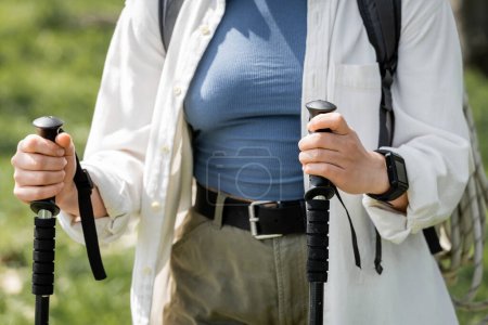 Cropped view of young woman hiker in casual clothes with backpack holding trekking poles while walking outdoors, independent traveler embarking on solo journey