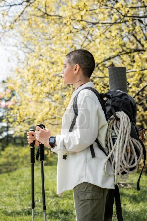 Photo for Side view of young short haired and tattooed female hiker with backpack and climbing rope holding trekking poles and walking on blurred nature, independent traveler embarking on solo journey - Royalty Free Image