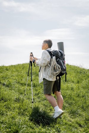 Side view of young short haired female traveler with backpack and climbing rope walking with trekking poles on hill with grass at background, independent traveler embarking on solo journey