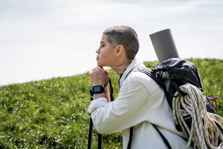 Photo for Side view of young short haired and tattooed young tourist with backpack holding trekking poles and looking away while standing with hill at background, explorer woman discovering hidden trails - Royalty Free Image