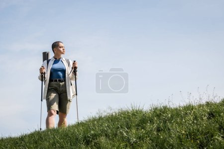 Junge kurzhaarige Wanderin in lässiger Kleidung mit Rucksack und Trekkingstöcken, die im Hintergrund auf einem grasbewachsenen Hügel und Himmel steht, Forscherin entdeckt versteckte Pfade