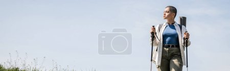 Junge kurzhaarige Reisende in Freizeitkleidung mit Rucksack, Trekkingstöcke in der Hand, im Hintergrund der Himmel, Forscherin entdeckt versteckte Pfade, Banner 