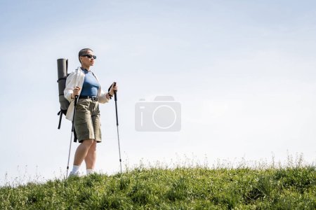 Viajero joven de pelo corto en gafas de sol con mochila y equipo de viaje sosteniendo bastones de trekking mientras camina en una colina cubierta de hierba, mujer exploradora descubriendo senderos ocultos