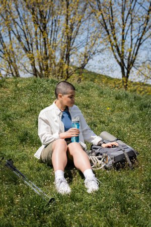 Young short haired female traveler holding sports bottle and sitting near backpack and trekking poles on grassy hill with nature at background, explorer woman, Translation of tattoo: love
