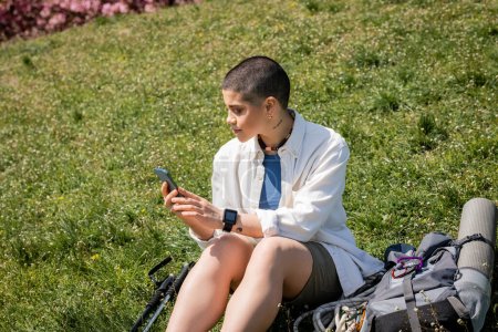 Young short haired and tattooed hiker in casual clothes using smartphone while sitting and relaxing near trekking poles and backpack on grassy hill, explorer woman discovering hidden trails