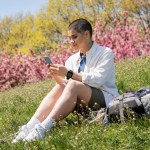 Smiling young short haired female traveler using smartphone while sitting near backpack with travel equipment on grassy hill with nature at background, curious hiker exploring new landscapes