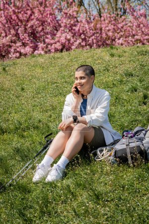Cheerful young short haired traveler talking on smartphone while sitting near trekking poles and backpack on grassy hill with nature at background, curious hiker exploring new landscapes