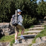 Young female hiker in baseball cap and casual clothes with backpack and travel equipment walking on stairs with nature and sky at background, curious hiker exploring new landscapes