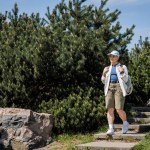 Young female hiker in casual clothes and baseball cap holding backpack while walking down on stairs with nature and blue sky at background, curious hiker exploring new landscapes