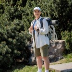 Positive young female traveler in baseball cap and casual clothes holding backpack and looking away while standing with nature at background, curious hiker exploring new landscapes