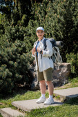 Positive young female traveler in baseball cap and casual clothes holding backpack and looking away while standing with nature at background, curious hiker exploring new landscapes tote bag #662549530