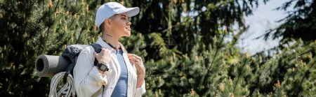 Photo for Side view of young and tattooed female tourist in casual clothes and baseball cap holding backpack while standing near blurred trees at background, curious hiker exploring new landscapes, banner - Royalty Free Image