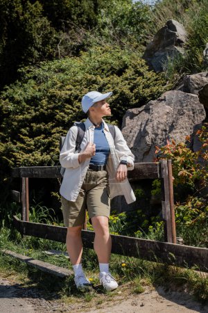 Side view of young short haired female traveler in casual clothes and baseball cap with backpack looking away while standing near fence outdoors, Translation of tattoo: love