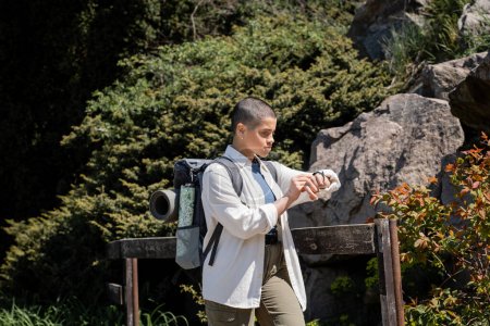Young short haired female traveler in casual clothes with backpack looking at wristwatch and standing near fence with nature at background, woman trekking across vast landscapes