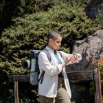 Young short haired female traveler in casual clothes with backpack looking at wristwatch and standing near fence with nature at background, woman trekking across vast landscapes