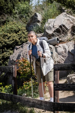 Young short haired woman traveler in casual clothes with backpack holding trekking poles and looking away while standing with nature at background, woman trekking across vast landscapes