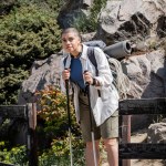 Young short haired woman traveler in casual clothes with backpack holding trekking poles and looking away while standing with nature at background, woman trekking across vast landscapes