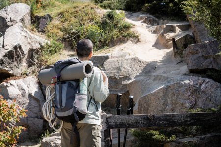 Rückenansicht einer jungen, kurzhaarigen Wanderin mit Rucksack, Fitnessmatte und Kletterseil im Hintergrund mit Hügel und Steinen, die durch weite Landschaften wandert
