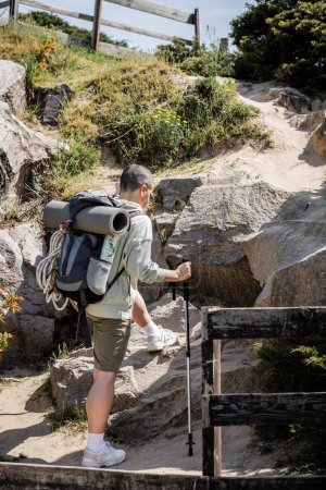 Junge kurzhaarige Touristinnen mit Rucksack und Trekkingstock, die im Sommer auf einem Hügel mit Steinen und der Natur im Hintergrund wandern, Frauen, die durch weite Landschaften wandern, Übersetzung von Tätowierung: Liebe