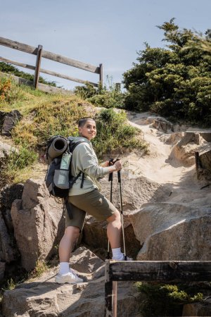 Positive junge kurzhaarige Wanderin mit Rucksack, die Trekkingstöcke in der Hand hält und im Sommer in der Nähe eines Hügels mit Steinen im Hintergrund steht, Übersetzung des Tattoos: Liebe